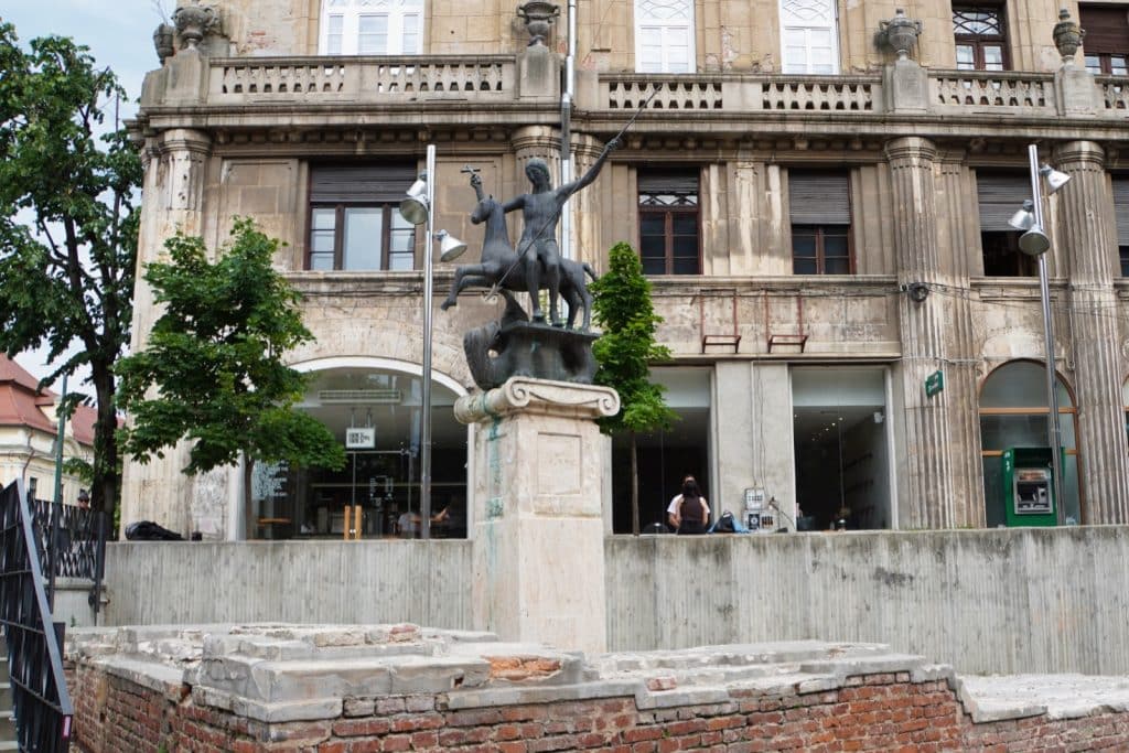 Sehenswürdigkeiten in Timișoara: Das Denkmal des Heiligen Georg in Timișoara zeigt eine Bronzestatue des Heiligen zu Pferd, der einen Drachen erschlägt, platziert auf einem hohen Steinsockel vor einem historischen Gebäude und archäologischen Ausgrabungen.
