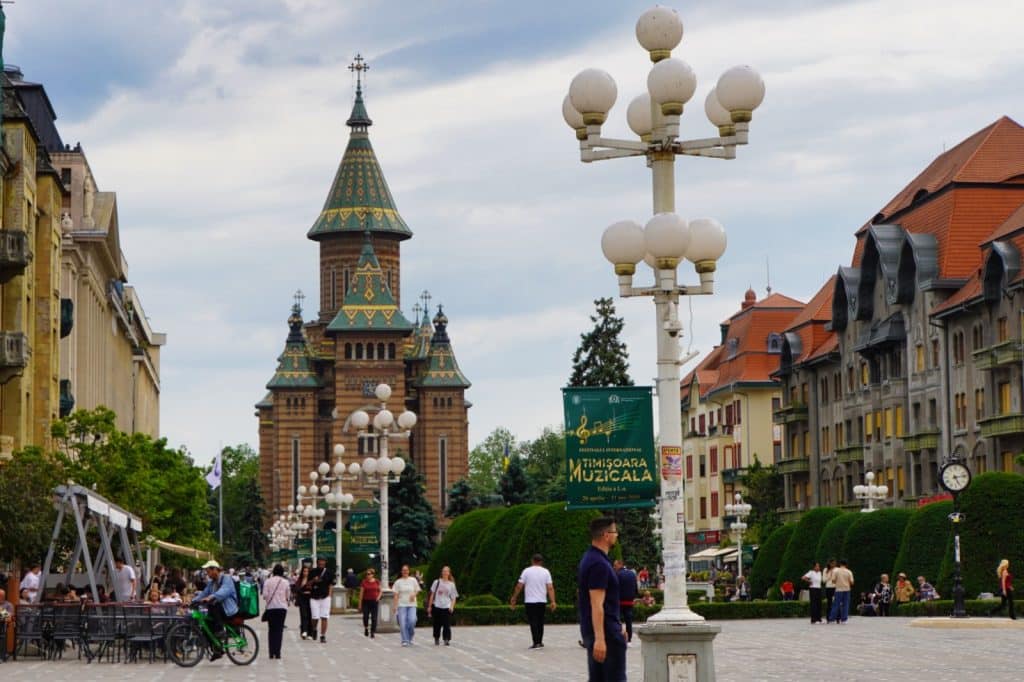 Blick entlang der belebten Flaniermeile in Timișoara auf die monumentale orthodoxe Kathedrale mit ihren charakteristischen bunt gemusterten Ziegeldächern unter einem bewölkten Himmel.