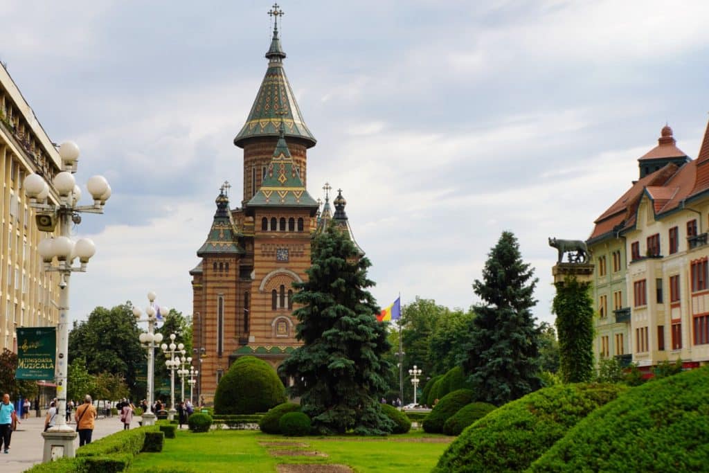 Blick über eine Grünanlage mit gepflegten Hecken auf die orthodoxe Kathedrale von Timișoara, deren markante Türme mit bunt glasierten Ziegeln gedeckt sind.