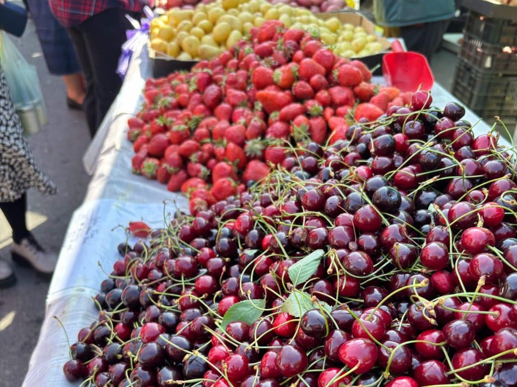 Ein Marktstand in Timișoara präsentiert große, aufgehäufte Mengen an frischen, dunkelroten Kirschen im Vordergrund sowie leuchtend rote Erdbeeren und gelbe Kartoffeln dahinter.
