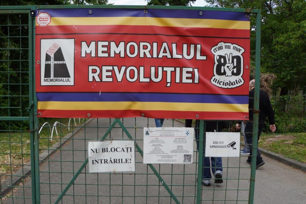 Ein rot-gelb-blaues Banner mit der Aufschrift „Memorialul Revoluției“ hängt am grünen Gittertor zum Eingang des Revolutionsmuseums in Timișoara. Darunter befinden sich Hinweisschilder in rumänischer Sprache, während im Hintergrund Besucher einen gepflasterten Weg entlanggehen.