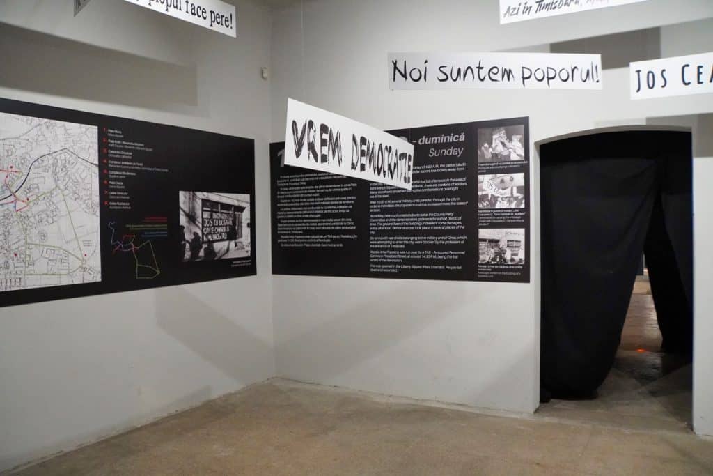 Ein Ausstellungsraum im Museum in Timișoara zeigt Informationstafeln mit Karten und historischen Fotos an den Wänden sowie von der Decke hängende Banner mit rumänischen Protestparolen wie „Vrem Democrație“. Die dokumentarische Installation beleuchtet die Ereignisse der rumänischen Revolution und führt zu einem mit schwarzen Vorhängen verhüllten Durchgang.