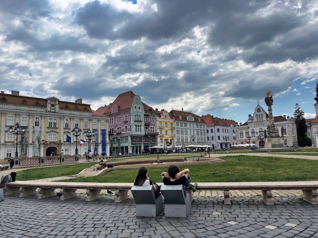 Zwei Personen sitzen auf modernen Metallstühlen am Rande des Union-Platzes (Piața Unirii) in Timișoara und blicken auf die historische Kulisse aus farbenfrohen Barockgebäuden unter einem dramatischen Wolkenhimmel.
