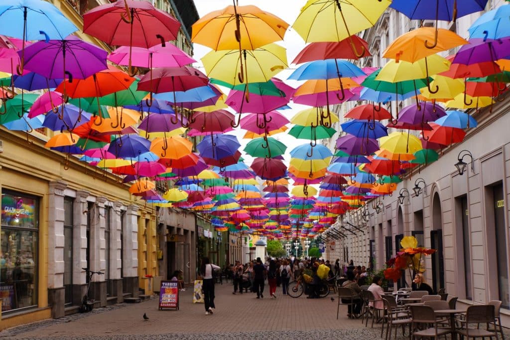 Blick entlang der belebten Regenschirmstraße in Timișoara, über der hunderte bunte Regenschirme in leuchtenden Farben wie Gelb, Rot, Blau und Violett als dekoratives Dach hängen. Passanten schlendern über das Kopfsteinpflaster der Fußgängerzone, die von historischen Gebäuden mit Straßencafés und Geschäften gesäumt wird.