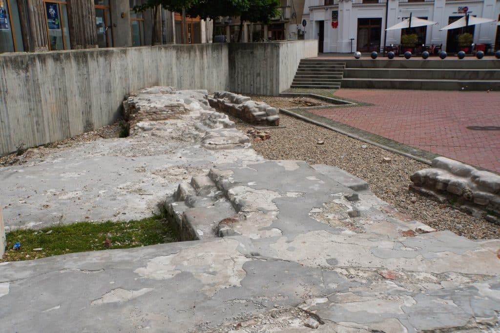 Sehenswürdigkeiten in Timișoara: Freigelegte steinerne Mauerreste und Fundamente einer historischen Kirche in Timișoara befinden sich in einer tiefergelegenen Ausgrabungsstätte, die von modernen Betonwänden und einem gepflasterten Platz umgeben ist.