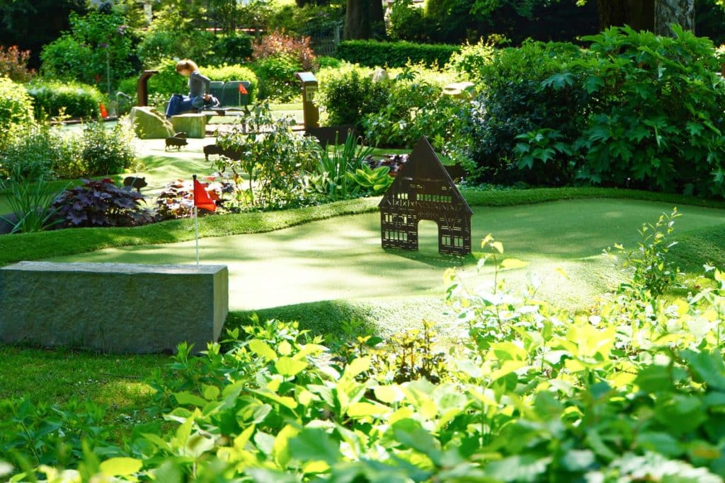 Ein sonniger Blick auf eine Minigolfanlage im Kurpark von Bad Sassendorf, bei der eine Spielbahn mit einem kleinen Modellhaus als Hindernis und einer roten Fahne im Vordergrund von üppigem Grün umgeben ist.