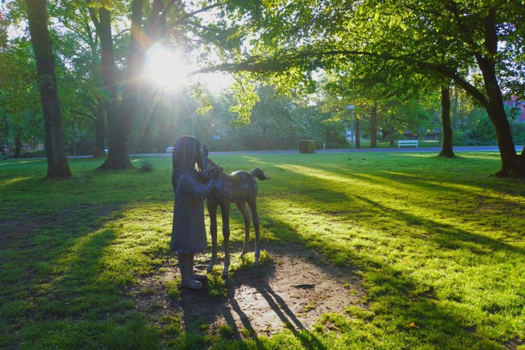 Eine Bronzestatue eines Mädchens, das ein Reh streichelt, steht im sonnendurchfluteten Kurpark von Bad Sassendorf auf einer Rasenfläche unter hohen Bäumen. Das warme Licht der tiefstehenden Sonne bricht durch das Laub und wirft lange Schatten auf das Gras.
