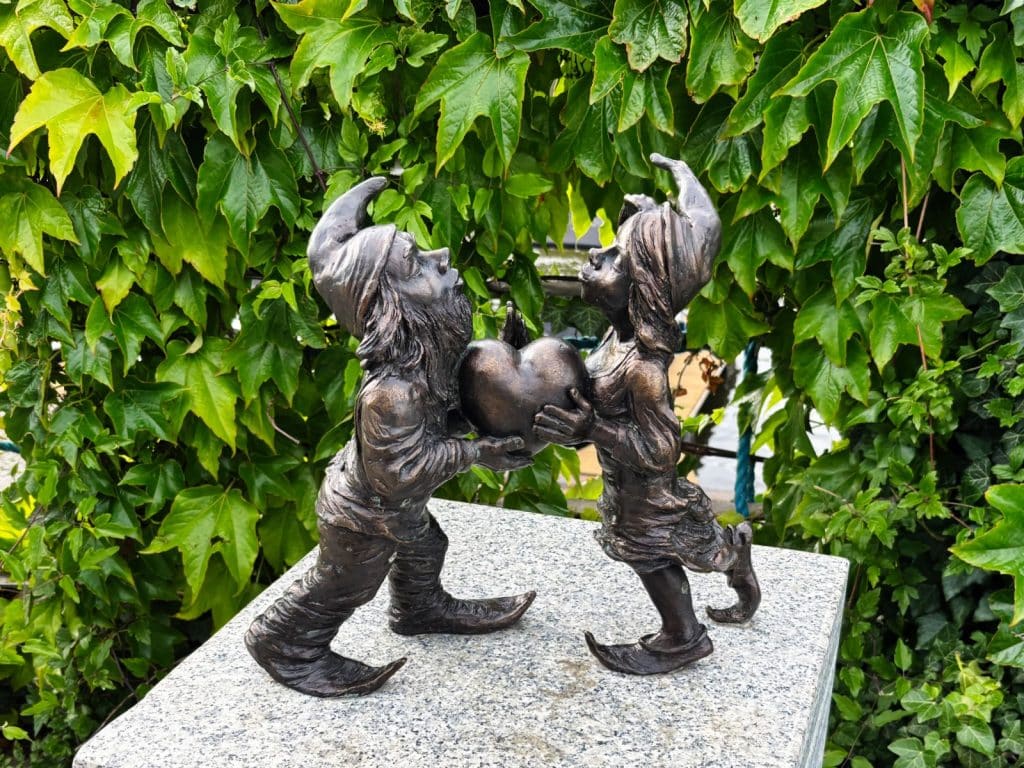 Zwei kleine Bronzefiguren der berühmten Breslauer Zwerge, ein männlicher und ein weiblicher Zwerg, stehen sich auf einem Steinsockel gegenüber und halten gemeinsam ein großes Herz in den Händen.