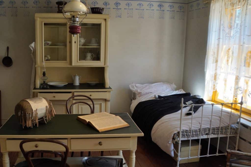 Ein historisch eingerichtetes Zimmer im Textilmuseum in Lodz mit einem weißen Metallbett, einem Schreibtisch mit aufgeschlagenem Buch und einem cremefarbenen Küchenbuffet vor einer gemusterten Tapete. Auf dem Tisch steht zudem ein Klöppelkissen zur Textilherstellung, während ein helles Fenster mit Spitzenvorhängen den Raum beleuchtet.