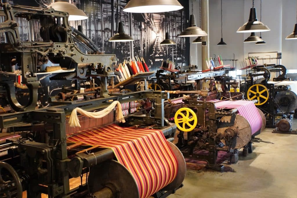 Historische Webstühle im Textilmuseum in Łódź weben bunt gestreifte Stoffe unter industriellen Hängelampen vor einer Wand mit einer großformatigen Schwarz-Weiß-Fotografie einer Fabrikhalle.