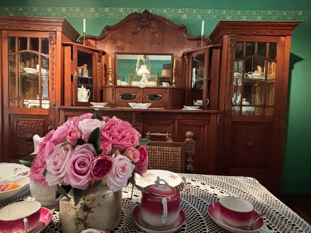 Ein detailreich gedeckter Kaffeetisch mit rosa Rosen und feinem Porzellan vor einem prunkvollen, dunklen Holzbuffet in einem historischen Wohnraum des Textilmuseums in Łódź. Das herrschaftliche Ambiente wird durch grüne Wände mit gemusterter Bordüre und eine weiße Häkeldecke unterstrichen.