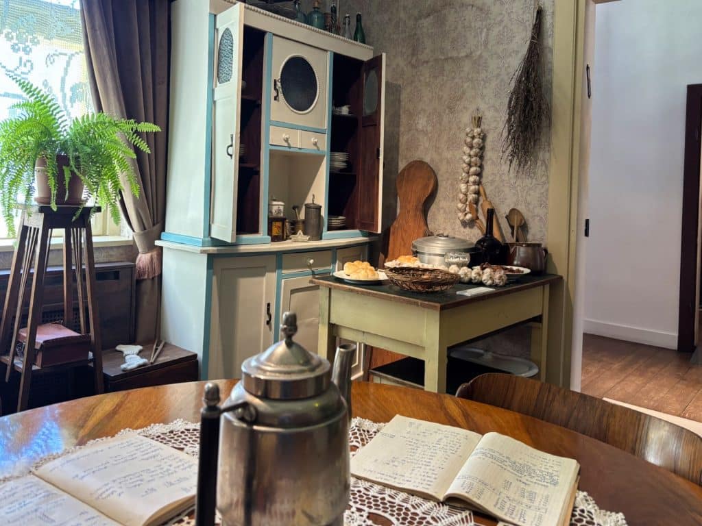 Ein historisch eingerichtetes Zimmer im Textilmuseum in Łódź mit einem runden Holztisch im Vordergrund, auf dem eine silberne Kanne und aufgeschlagene Hefte liegen, sowie einem hellblauen Küchenschrank und einem Küchentisch mit Lebensmitteln im Hintergrund.