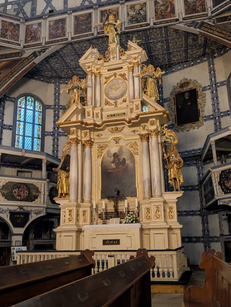 Der prunkvolle, mehrstöckige Barockaltar der Friedenskirche in Jawor, Niederschlesien, ist mit goldenen Skulpturen, Marmorsäulen und religiösen Gemälden vor einer blau-weißen Fachwerkwand reich verziert. Im Vordergrund stehen hölzerne Kirchenbänke, während die Decke und Emporen mit zahlreichen biblischen Malereien geschmückt sind.