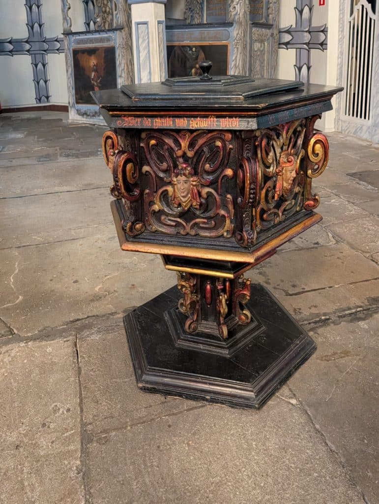 Ein reich verziertes, hölzernes Taufbecken aus der Friedenskirche in Jawor, Niederschlesien, mit barocken Schnitzereien, Engelsköpfen und einer deutschen Inschrift auf einem sechseckigen Sockel.