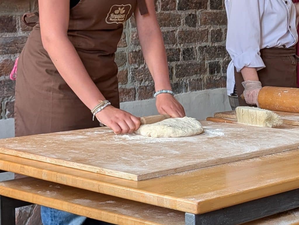 Eine Person in einer braunen Schürze rollt auf einem bemehlten Holzbrett Teig für Posener Martinsbrot aus, während im Hintergrund eine weitere Person mit einem Nudelholz bereitsteht.