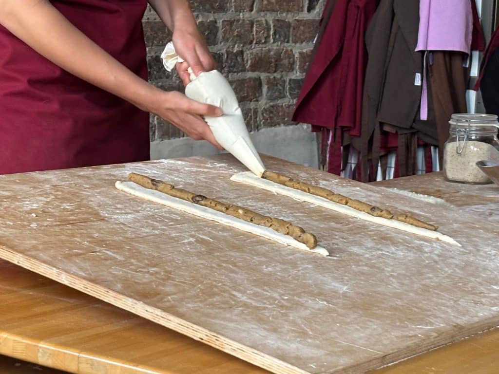 Eine Person in einer dunkelroten Schürze spritzt mit einem Spritzbeutel eine braune Füllung auf zwei lange Teigstreifen, um Martinshörnchen vorzubereiten.