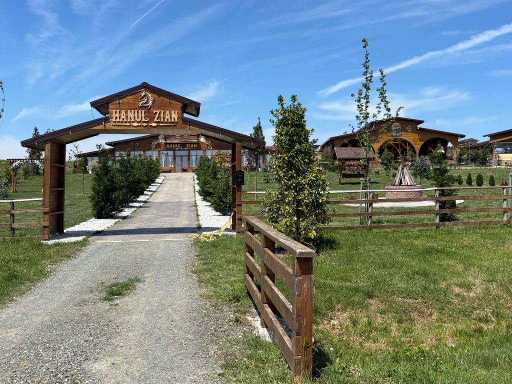 Ein hölzerner Torbogen mit der Aufschrift „Hanul Zian“ markiert den Eingang zum Grillrestaurant in der Region Timiș, das von weitläufigen Grünflächen und rustikalen Holzzäunen umgeben ist. Ein befestigter Weg führt unter strahlend blauem Himmel direkt auf das Hauptgebäude im Hintergrund zu.