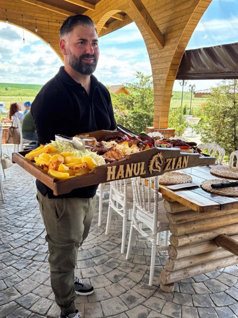 Ein lächelnder Mann serviert in einem Grillrestaurant in der Region Timiș eine riesige, rustikale Holzplatte mit verschiedenen Fleischsorten, Pommes frites und Beilagen, auf der der Schriftzug „HANUL ZIAN“ eingraviert ist.