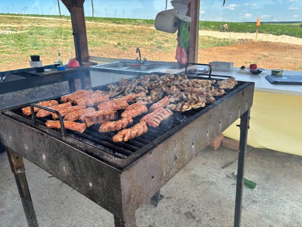 Ein großer, rustikaler Metallgrill im Grillrestaurant Hanul Zian in der Region Timis ist dicht mit verschiedenen Fleischsorten wie Mici und Hähnchenstücken belegt, die über glühender Kohle garen.