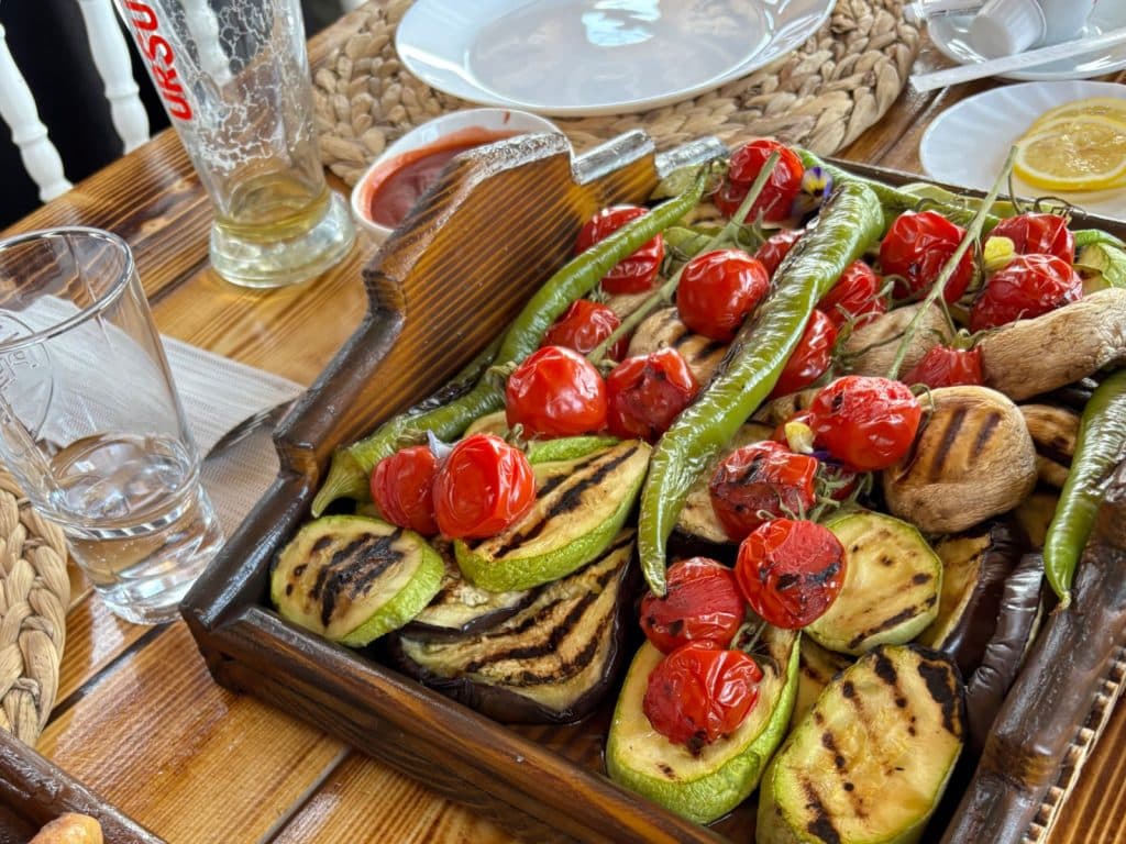 Eine rustikale Holzplatte mit gegrilltem Gemüse, darunter Zucchini, Auberginen, Champignons, Kirschtomaten und grüne Peperoni, serviert in einem Grillrestaurant in der Region Timis.