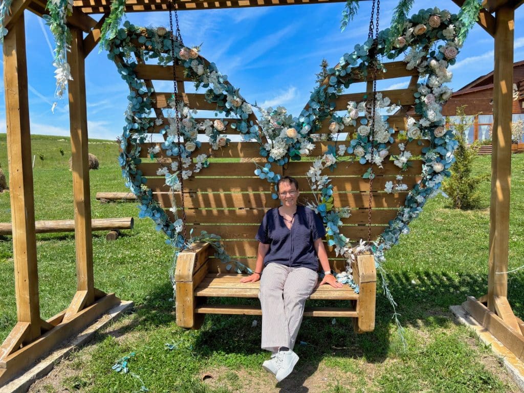 Eine Frau sitzt lächelnd auf einer hölzernen Schaukel in Schmetterlingsform, die mit blau-weißen Blumen dekoriert ist, im Außenbereich des Hanul Zian.