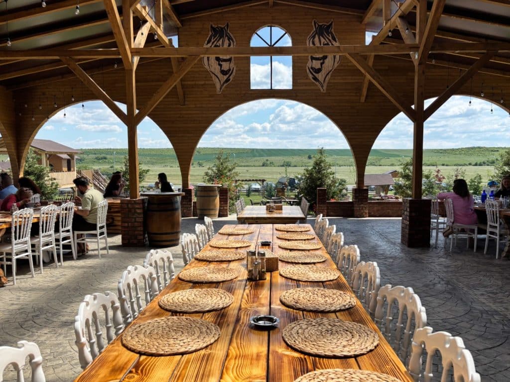 Die überdachte Holzterrasse eines Grillrestaurants in der Region Timiș bietet einen weiten Blick auf grüne Felder durch große Rundbögen, während im Vordergrund eine lange Tafel mit runden Platzsets aus Korbgeflecht und weißen Stühlen für Gäste bereitsteht.