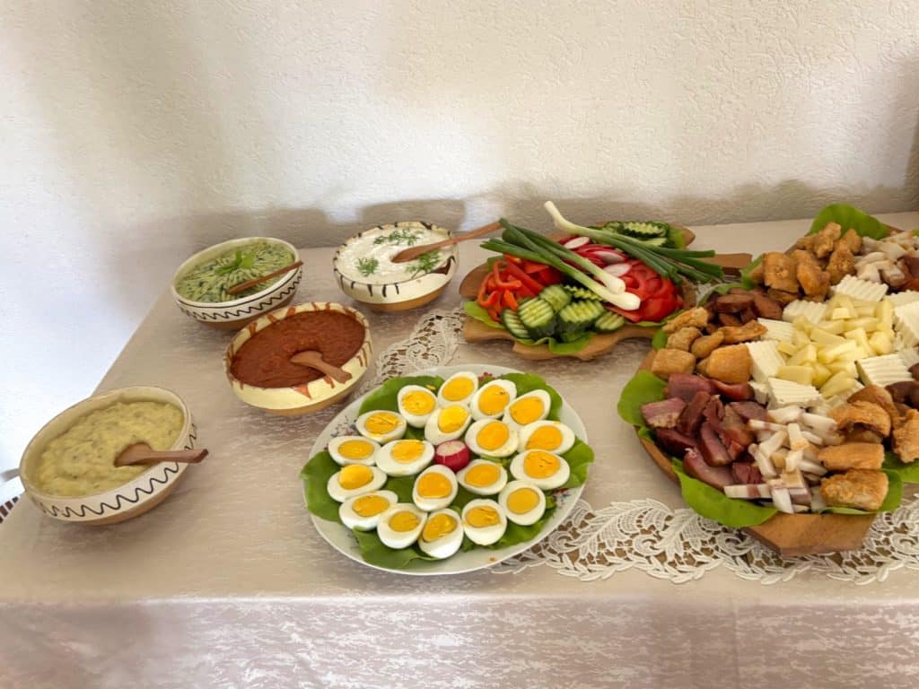 Ein reichhaltig gedeckter Tisch in der Region Timis zeigt verschiedene Schüsseln mit Aufstrichen, eine Platte mit halbierten gekochten Eiern, frisches Gemüse wie Gurken und Radieschen sowie eine große Holzplatte mit Speck, Käsewürfeln und Grieben.