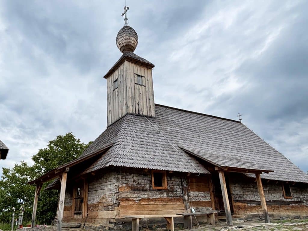 Eine historische Holzkirche in der Region Timiș mit einem schindelgedeckten Dach, einem markanten Glockenturm und einer kleinen Zwiebelkuppel unter einem bewölkten Himmel.
