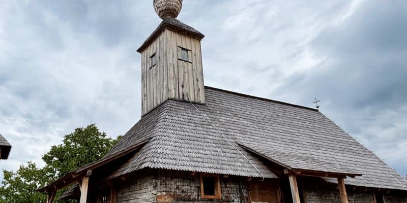 Eine historische Holzkirche in der Region Timiș mit einem schindelgedeckten Dach, einem markanten Glockenturm und einer kleinen Zwiebelkuppel unter einem bewölkten Himmel.