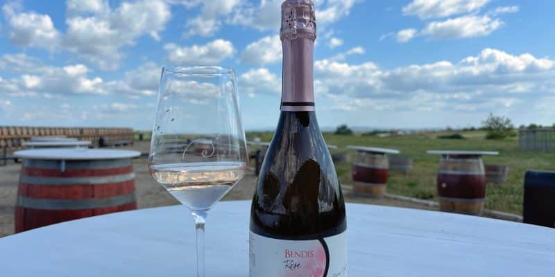 Eine Flasche Bendis Rosé Schaumwein steht neben einem gefüllten Weinglas auf einem weißen Tisch vor der Kulisse eines Weinguts in der Region Timiș unter blauem Himmel.
