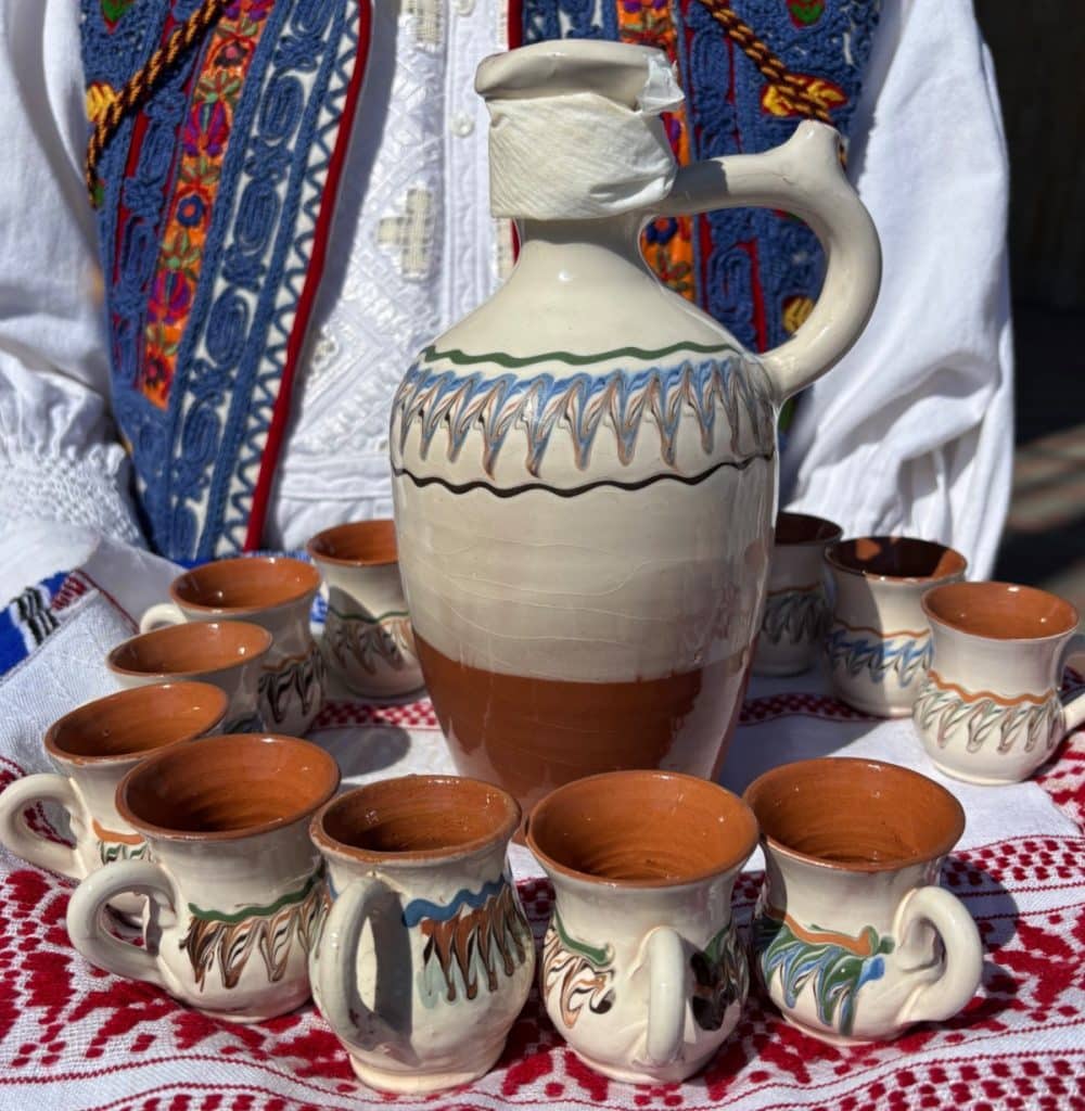 Eine dekorative rumänische Keramikkanne für Schnaps steht umringt von elf passenden kleinen Bechern auf einem rot-weiß bestickten Tuch vor einer Person in traditioneller Tracht.