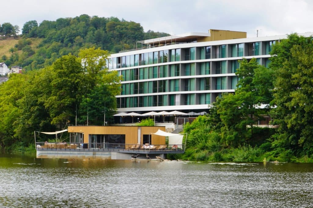 Blick über die Lahn auf die moderne Flusssauna der Emser Therme in Bad Ems, die als hölzerner Pavillon mit Sonnensegeln auf dem Wasser schwimmt, dahinter das mehrstöckige Hotelgebäude.