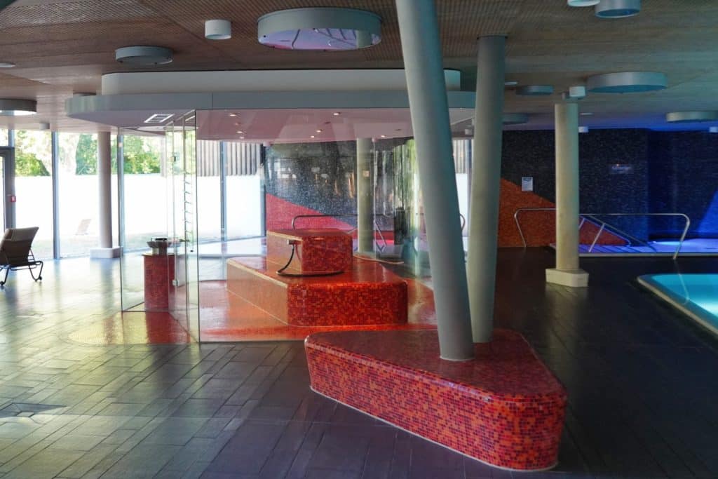 Das Kräuterbad der Emser Therme in Bad Ems zeigt einen modernen Wellnessbereich mit roten Mosaik-Sitzbänken, einer gläsernen Dampfkabine und schrägen Stützsäulen vor einem dunklen Fliesenboden.