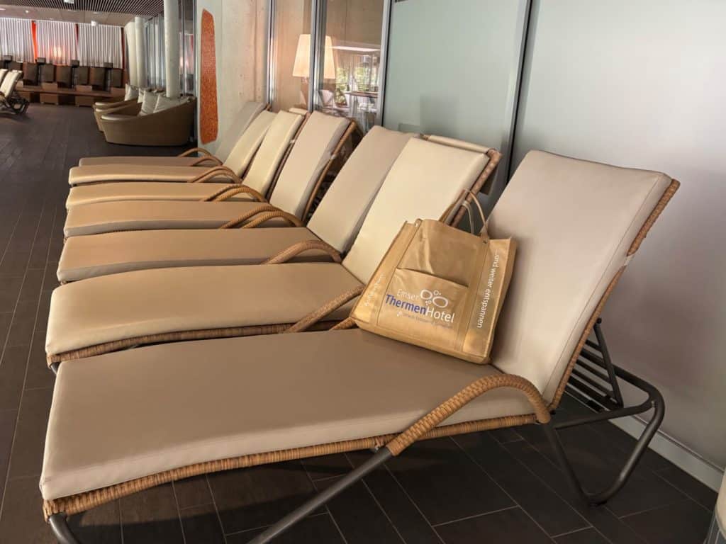 In der Liegenlandschaft der Emser Therme in Bad Ems stehen mehrere beige gepolsterte Ruheliegen ordentlich nebeneinander, wobei auf der vordersten Liege eine braune Tasche mit dem Logo des ThermenHotels platziert ist.