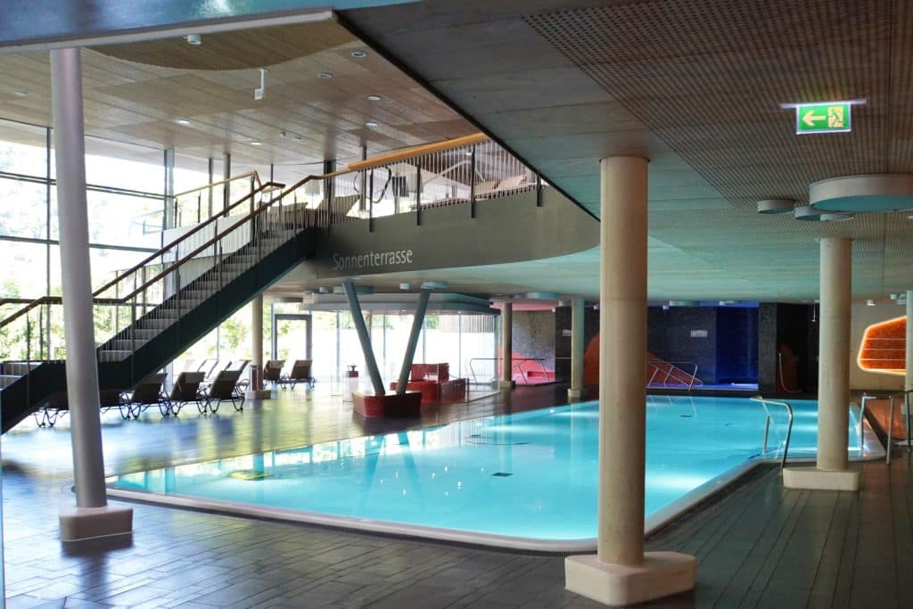 Innenansicht der Emser Therme in Bad Ems mit einem großen, hellblau leuchtenden Thermalbecken, das von modernen Säulen, einer Treppe zur Sonnenterrasse und Liegestühlen umgeben ist.