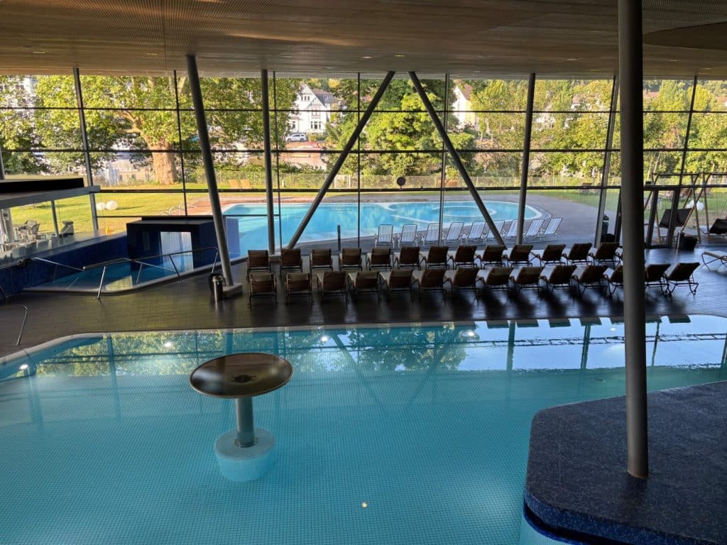Blick von oben auf die Innen- und Außenbecken der Emser Therme in Bad Ems mit zahlreichen Liegestühlen vor einer großen Glasfront.
