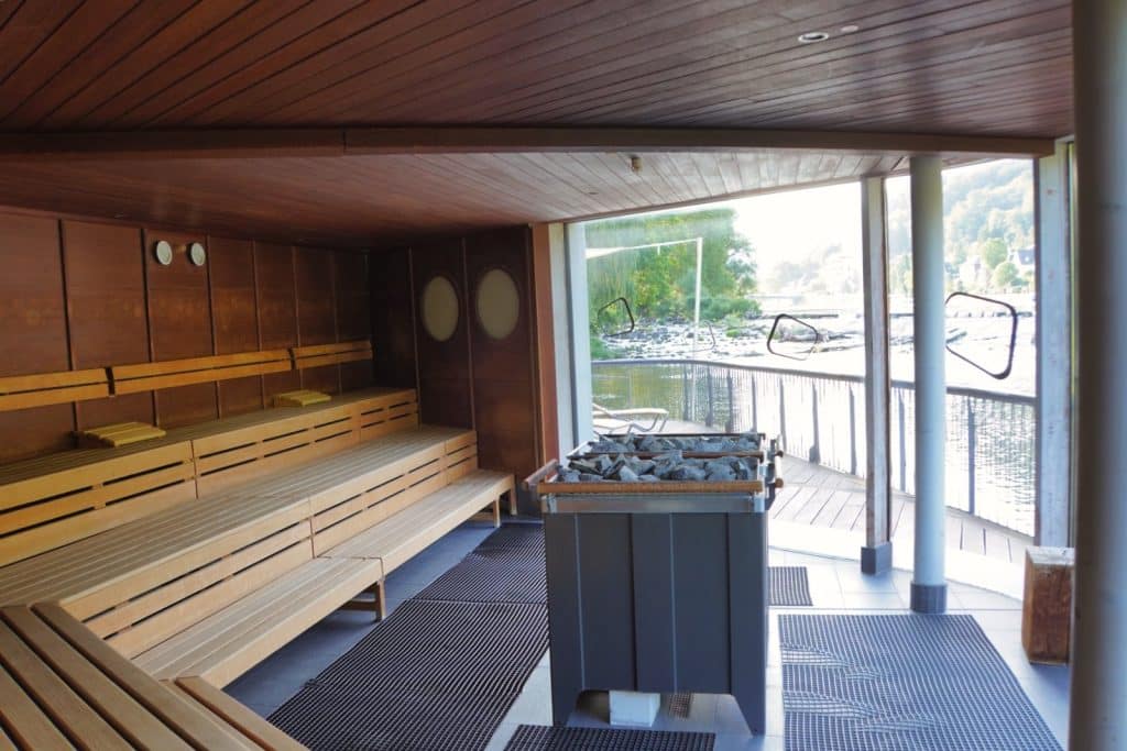 Innenansicht der Flusssauna in der Emser Therme mit hölzernen Sitzbänken auf mehreren Ebenen, einem zentralen Saunaofen mit Steinen und einem großen Panoramafenster mit Blick auf die Lahn.