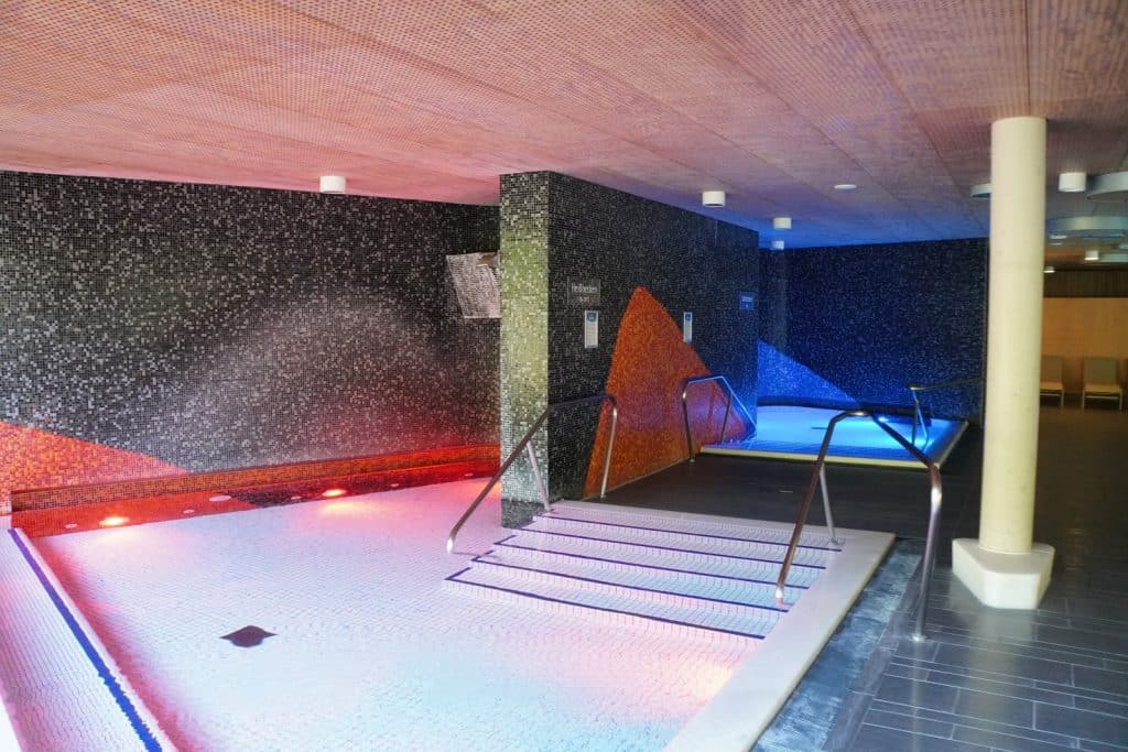 Innenansicht der Emser Therme mit einem Heißbecken in rötlichem Licht und einem Kaltbecken in blauem Licht, die durch Mosaikwände und Metallgeländer getrennt sind.