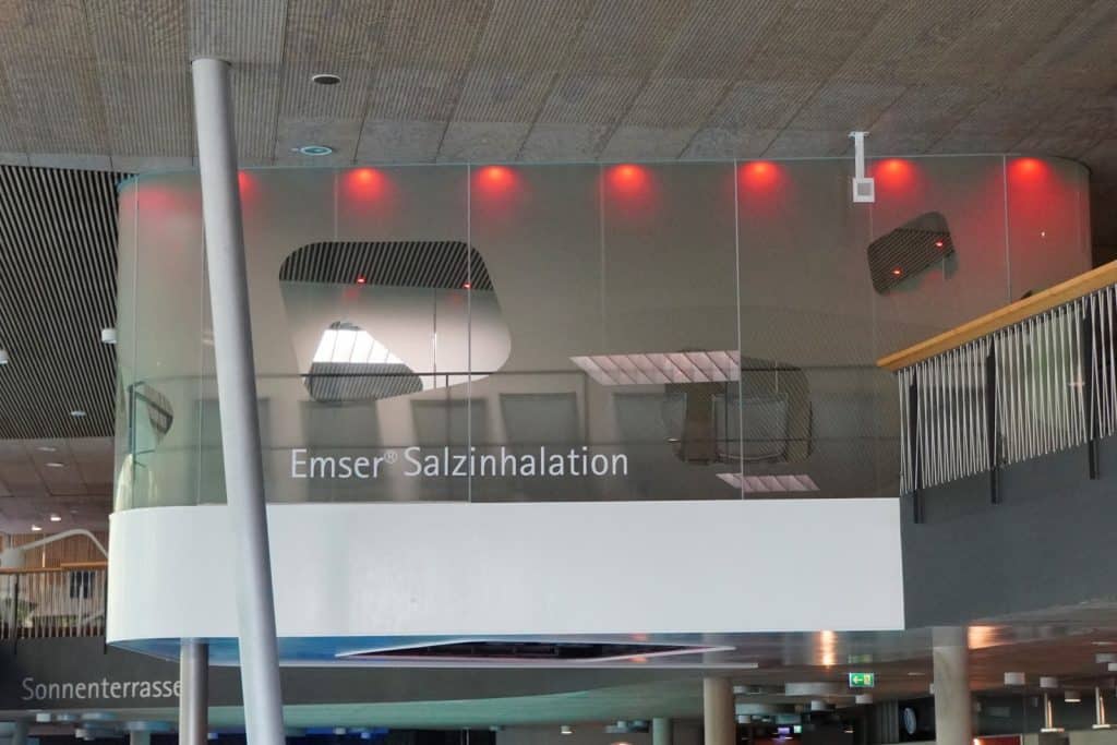 Außenansicht des Inhalationsraums der Emser Therme in Bad Ems mit einer Glasfront, auf der der Schriftzug „Emser Salzinhalation“ steht und die durch rote Lichter oberhalb beleuchtet wird.