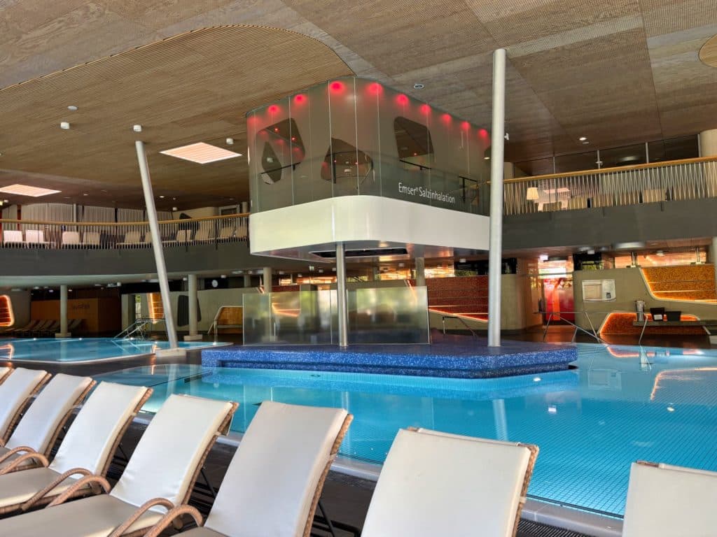 Innenansicht der Emser Therme mit einer Reihe weißer Liegen im Vordergrund, die auf ein hellblaues Thermalbecken und eine zentrale Emser Salzinhalation-Kabine blicken.