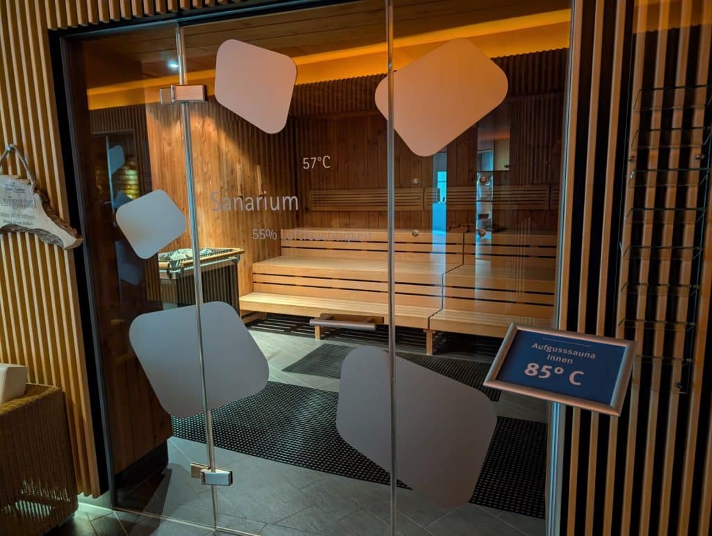 Blick durch eine Glastür in das Sanarium der Bad Ems Therme mit Holzbänken, einer Temperaturanzeige von 57 °C und 55 % Luftfeuchtigkeit.