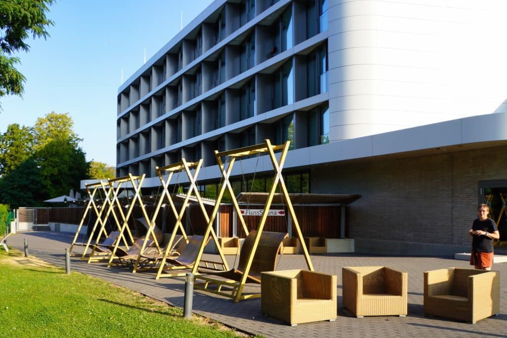 Außenbereich der Emser Therme in Bad Ems mit einer Reihe hölzerner Schaukelsessel und beigen Korbsesseln vor einem modernen, mehrstöckigen Hotelgebäude.