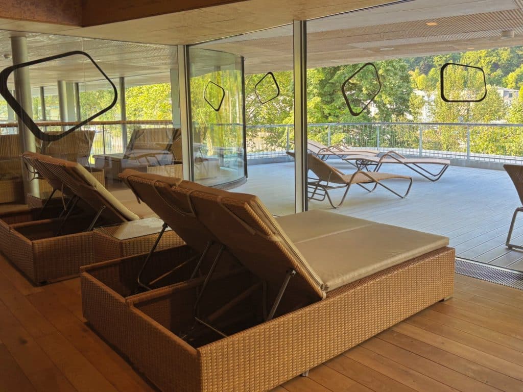 Die obere Sonnenterrasse der Emser Therme in Bad Ems bietet einen Entspannungsbereich mit gepolsterten Rattan-Liegen im Innenraum und weiteren Liegestühlen auf dem angrenzenden Außendeck mit Blick ins Grüne.