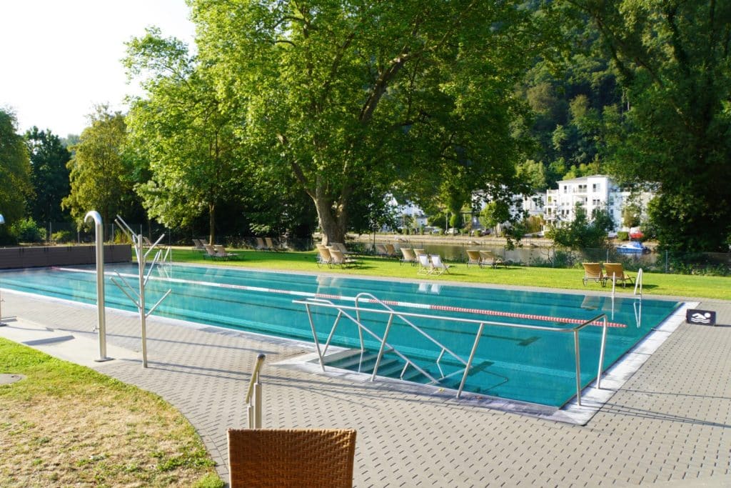 Das Sportbecken der Emser Therme in Bad Ems mit türkisblauem Wasser, Edelstahltreppen und Schwimmleinen, umgeben von einer Liegewiese mit Sonnenstühlen und hohen Bäumen direkt an der Lahn.
