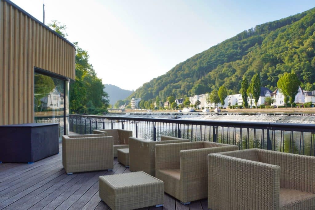 Blick von der Terrasse der Flusssauna der Bad Ems Therme auf eine Sitzgruppe aus Korbgeflecht, die direkt an der Lahn mit Aussicht auf bewaldete Hügel und Wohnhäuser liegt.
