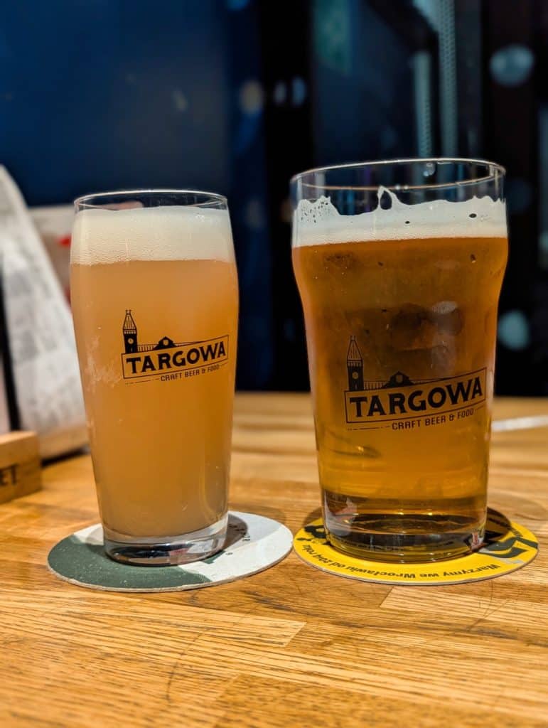 Zwei Gläser Bier der Marke Targowa stehen auf einem Holztisch in Breslau, wobei das linke Glas ein trübes, rosafarbenes Litovel Pink Guava und das rechte ein klares, goldenes Bier enthält.