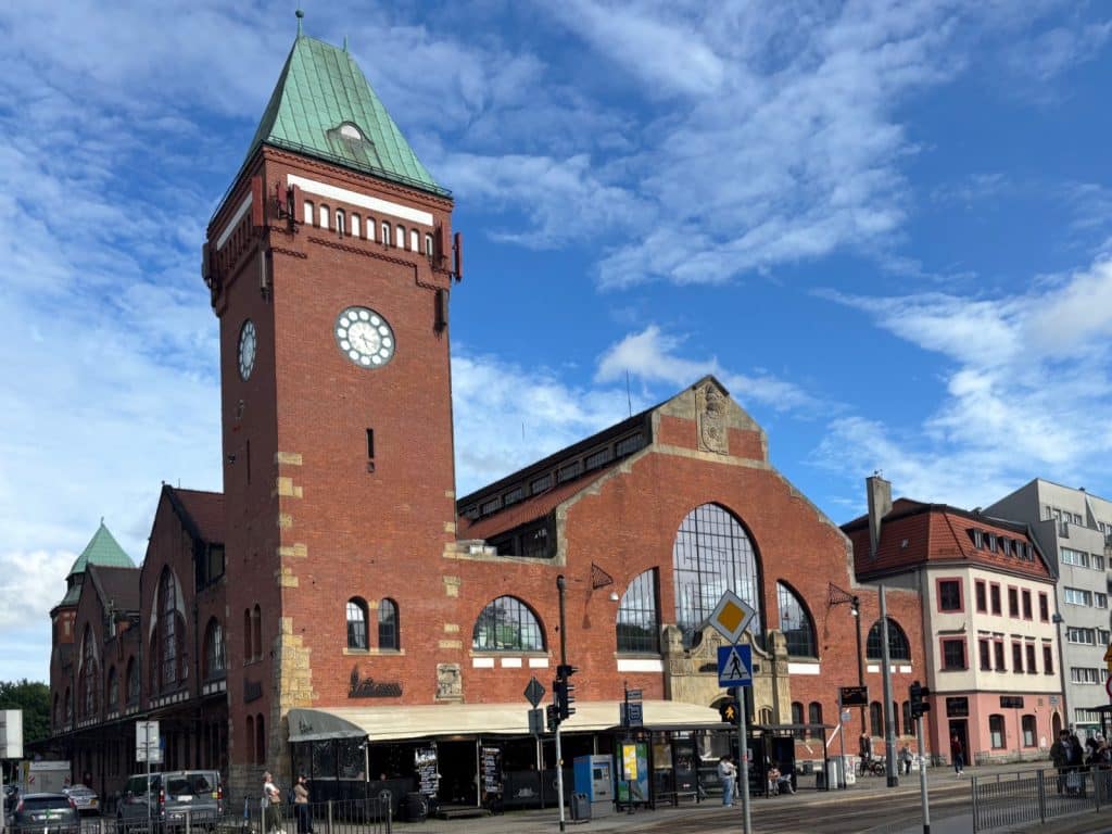 Die historische Markthalle (Hala Targowa) in Breslau aus rotem Backstein mit einem markanten Uhrturm und grünen Kupferdächern unter einem leicht bewölkten blauen Himmel. Vor dem Gebäude befinden sich eine Außengastronomie, eine Straßenbahnhaltestelle und Passanten im städtischen Umfeld.