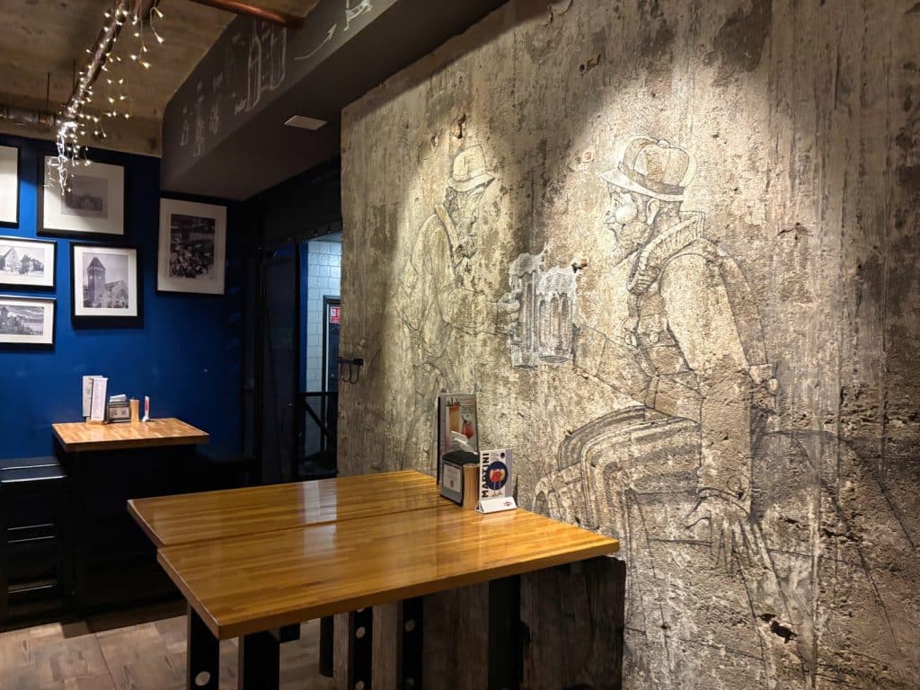 Innenansicht des Gastraums von Targowa in Breslau mit Holztischen vor einer rustikalen Betonwand, auf der eine große Zeichnung von zwei Männern beim Biertrinken zu sehen ist.