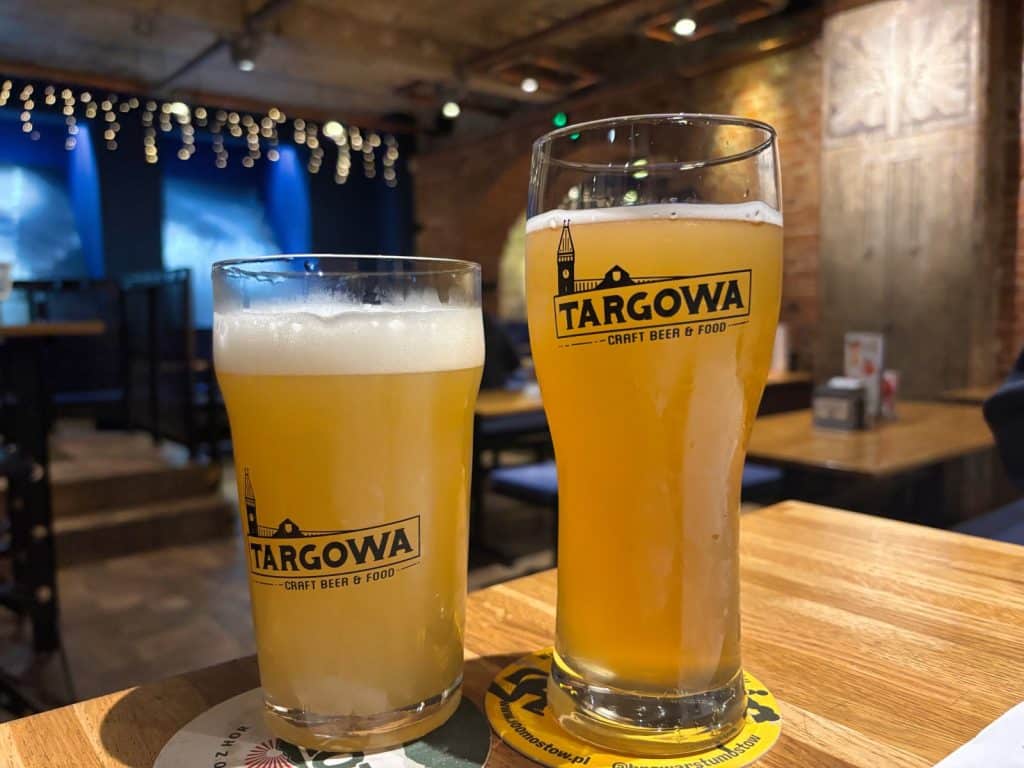 Zwei Gläser helles, trübes „Sandy Tower Sour“-Bier stehen auf einem Holztisch im Restaurant Targowa in Breslau, vor einem stimmungsvoll beleuchteten Hintergrund mit Ziegelwänden.