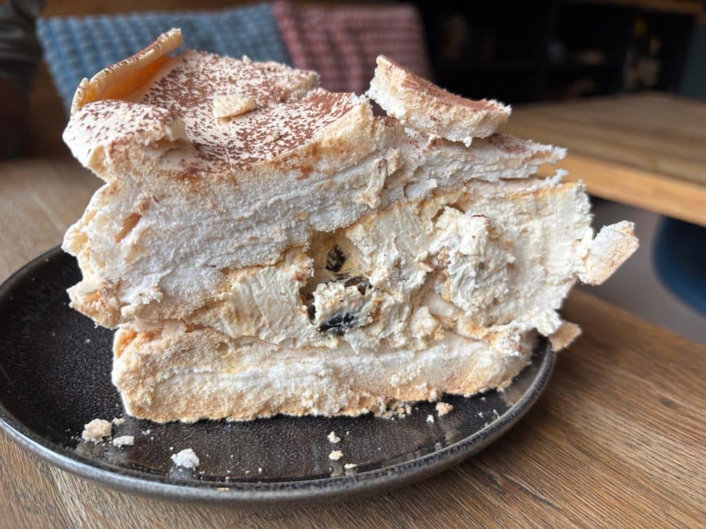 Ein großes Stück Baiserkuchen aus dem Lodz Bistro Montag mit cremiger Füllung und dunklen Beeren auf einem dunklen Teller.